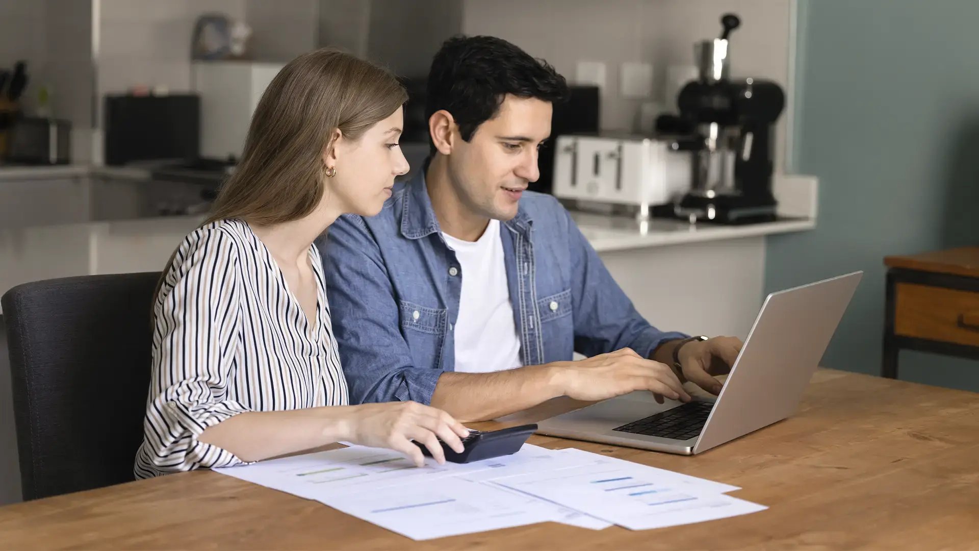 Pareja joven revisa sus finanzas con una calculadora, documentos y un notebook en la mesa de su casa.